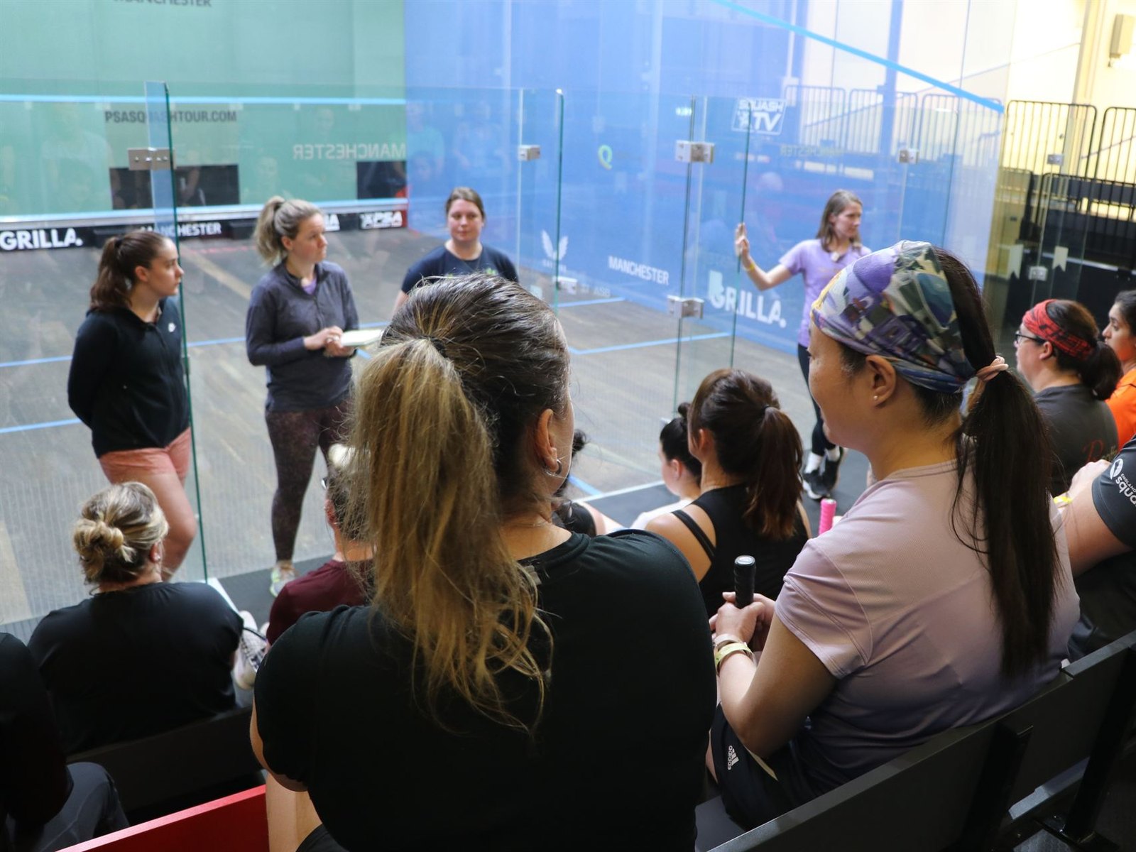 A coach briefing a group of players beside a glass squash court